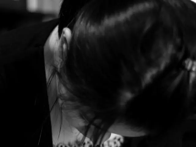 In the image a woman is sitting and holding a pen and writing and there is a book.