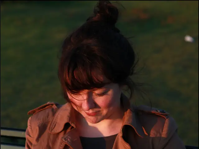 There is a lady wearing jacket is sitting on a bench. In the background it is green and blurred.