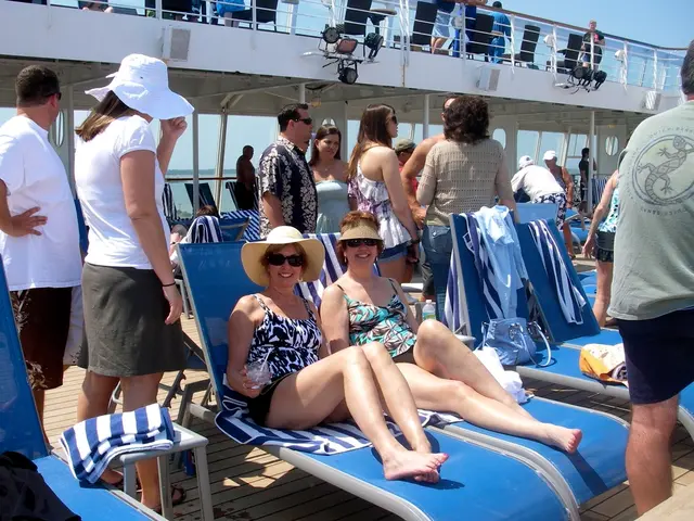 In the image there are two women in bikini sitting on chairs and behind them there are many people...
