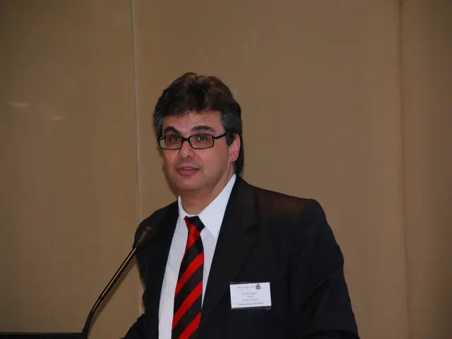 At the bottom of this image, there is a person in a suit, wearing a badge and a spectacle, smiling...