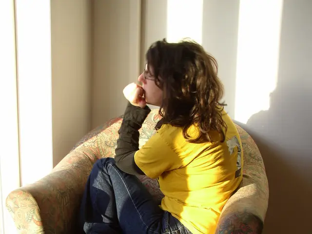 In a picture a person is sitting on a chair with a cushion on it there is a wall near to him.