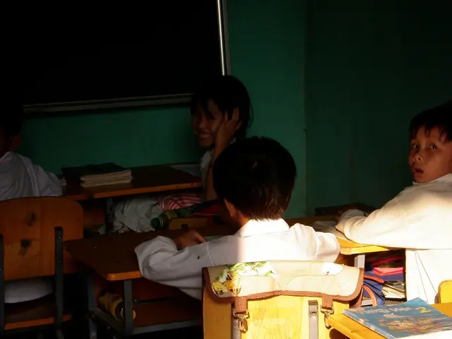 This children are sitting on a chairs. On this tables there are books. A blackboard on wall.